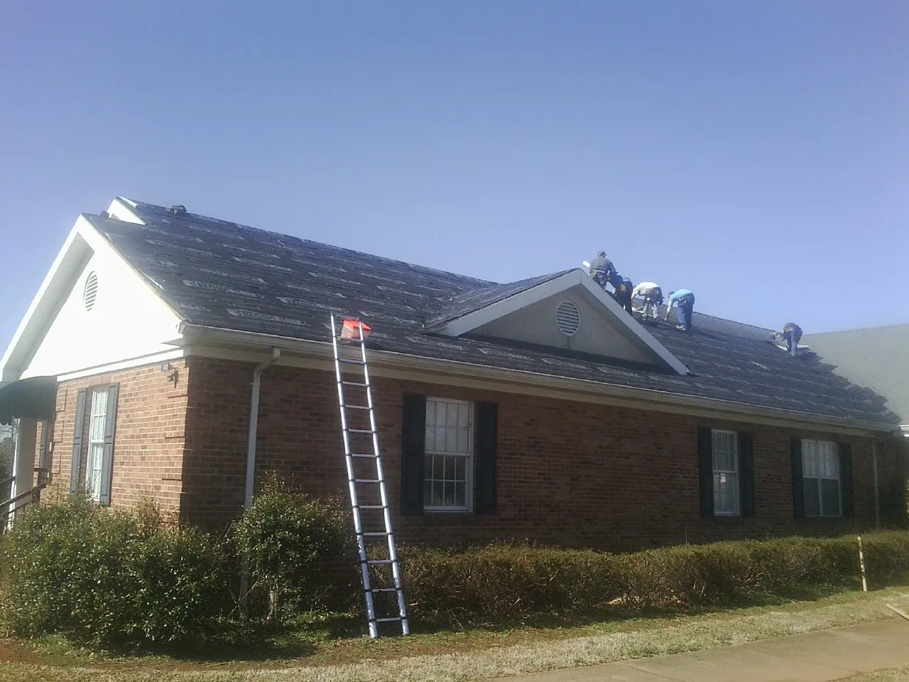 Crew tearing shingles off a brick ranch roof with a ladder set — Metal Roof Repair work by our crew in Finley