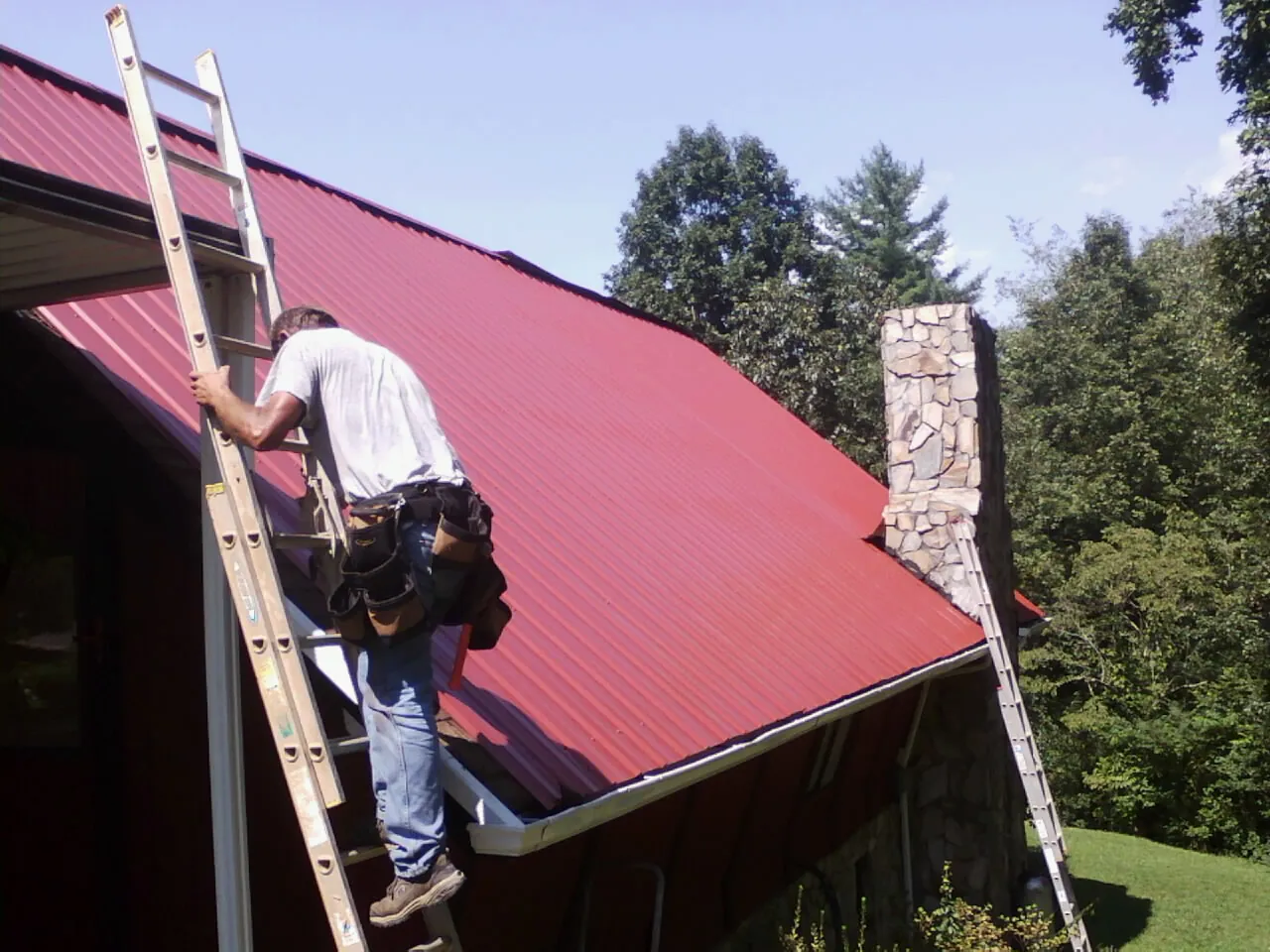 Roofer on a ladder beside a red metal roof and stone chimney — Soffit & Fascia Repair by Roofing Brothers in Finley