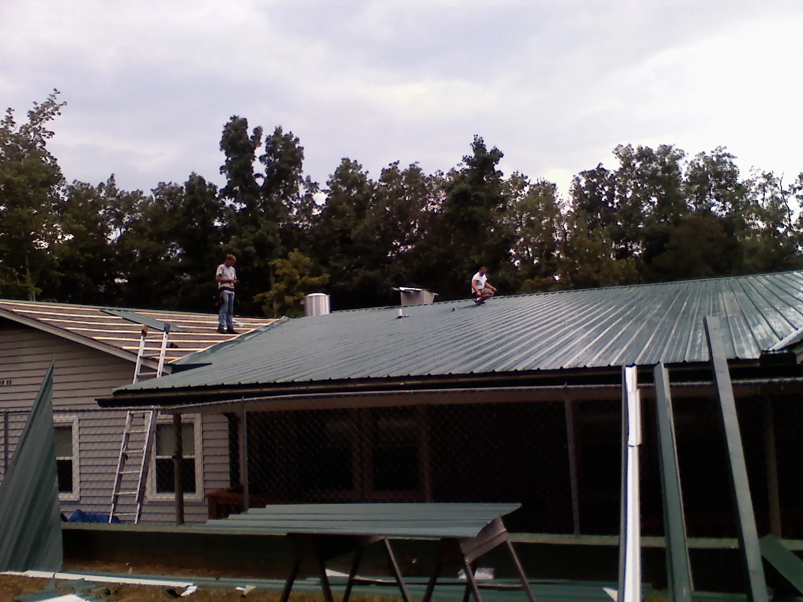 Two roofers installing green metal panels above a covered porch in Finley